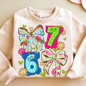 May include: A beige sweatshirt featuring the numbers 6 and 7, along with two floral bows and two additional bows with floral patterns. The design includes hearts and flowers in various colors.