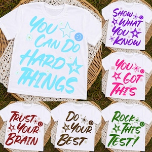 Peut inclure: T-shirts blancs avec des phrases motivantes de différentes couleurs. Les phrases incluent « You Can Do Hard Things », « Show What You Know », « You Got This », « Trust Your Brain », « Do Your Best » et « Rock This Test ! » Les motifs comportent des étoiles et des smileys.