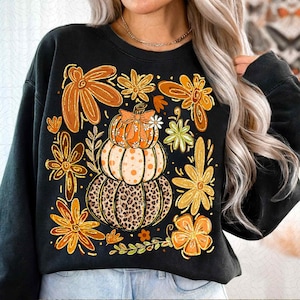 May include: Black sweatshirt featuring a stacked pumpkin design with a leopard print base, a dotted middle, and an orange top. The design is surrounded by orange and yellow flowers and green leaves, perfect for fall.