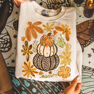 May include: Beige sweatshirt featuring a stacked pumpkin design with a leopard print base, polka dots, and a bow. Surrounding the pumpkin are orange and gold floral accents, perfect for autumn. The sweatshirt is held by hands.