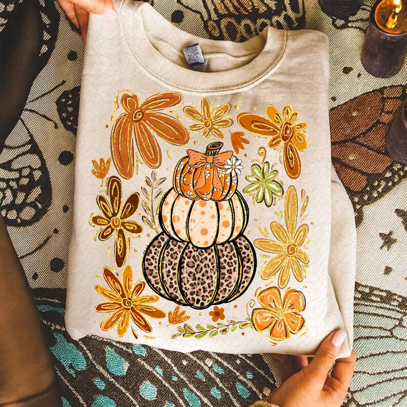 May include: Beige sweatshirt featuring a stacked pumpkin design with a leopard print base, polka dots, and a bow. Surrounding the pumpkin are orange and gold floral accents, perfect for autumn. The sweatshirt is held by hands.
