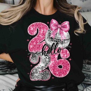 May include: Black t-shirt with a sparkly pink "26" design and a disco ball with a pink bow. The word "hello" is written in cursive across the design. The design is embellished with pink star accents.