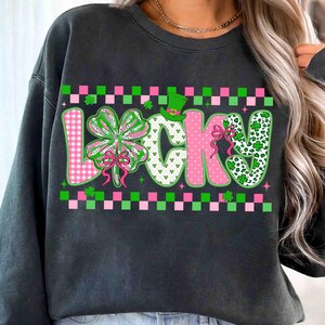 May include: Dark gray sweatshirt with the word "LUCKY" in a colorful design. The "L" is pink gingham, the "U" is white with shamrocks, the "C" is pink polka dots, the "K" is pink with a bow, and the "Y" is white with shamrocks. A green leprechaun hat sits above the "C".