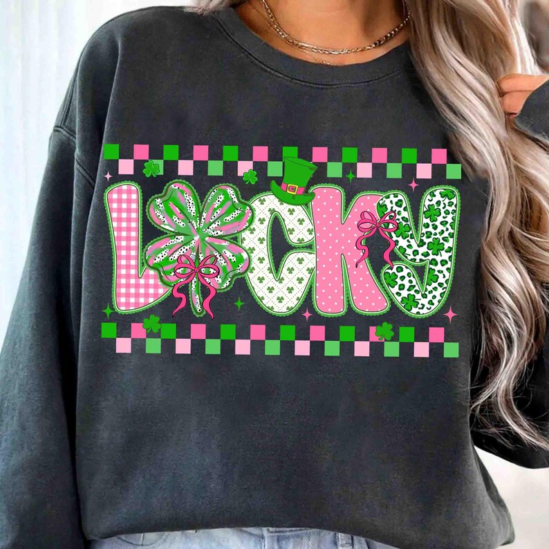 May include: Dark gray sweatshirt with the word "LUCKY" in a colorful design. The "L" is pink gingham, the "U" is white with shamrocks, the "C" is pink polka dots, the "K" is pink with a bow, and the "Y" is white with shamrocks. A green leprechaun hat sits above the "C".