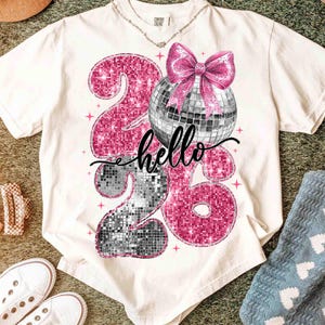 May include: Cream-colored t-shirt with a graphic design featuring the numbers "26" in pink and silver glitter, a disco ball, and a pink bow. The word "hello" is written in cursive. A necklace is visible.