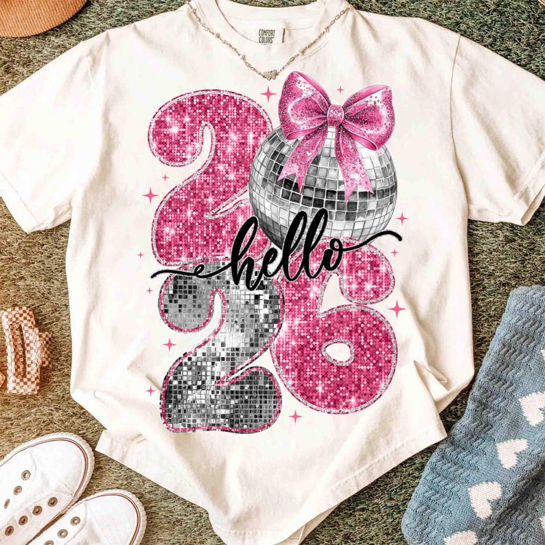 May include: Cream-colored t-shirt with a graphic design featuring the numbers "26" in pink and silver glitter, a disco ball, and a pink bow. The word "hello" is written in cursive. A necklace is visible.
