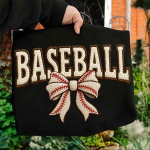以下が含まれることがあります： 黒色のトートバッグ。白い「BASEBALL」の文字が茶色で縁取られ、野球のリボンのデザインが施されています。リボンは白で、赤いステッチが施されています。バッグは人が持っています。