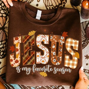 May include: A brown sweatshirt with the words "JESUS is my favorite season" in a decorative font. The letters are filled with fall-themed patterns, including pumpkins, plaid, and floral designs. Autumn leaves are scattered around the text.