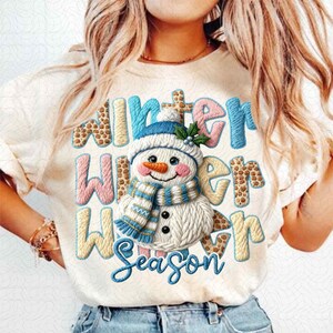 May include: Cream-colored t-shirt featuring a snowman design and the words "Winter Winter Season" in a mix of blue, pink, and tan colors. The snowman wears a blue hat and scarf.