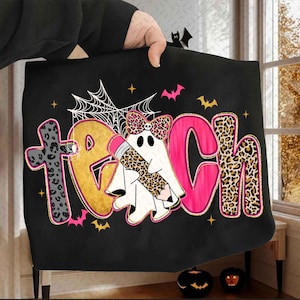 Peut inclure: Un sweat-shirt noir avec le mot TEACH &eacute;crit en lettres color&eacute;es et &agrave; motifs. La lettre A est remplac&eacute;e par un fant&ocirc;me blanc portant un n&oelig;ud &agrave; motif l&eacute;opard et tenant un crayon. Le motif inclut une toile d'araign&eacute;e, des chauves-souris et des &eacute;toiles.