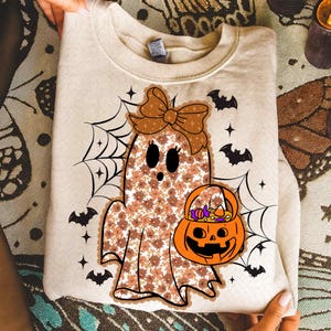 May include: Beige Halloween sweatshirt with a floral ghost design, bow, and jack-o'-lantern candy bucket. Black bats, spiderwebs, and stars complete the design. The sweatshirt is folded.