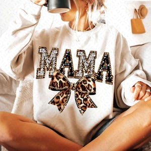 May include: Cream-colored sweatshirt with the word "MAMA" in a jeweled, black and white design. A leopard print bow is centered below the word. The sweatshirt is a casual, comfortable style.