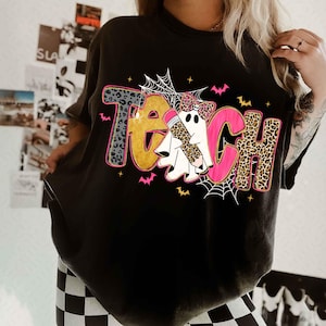 May include: Black t-shirt with the word "TEACH" in a playful design. The letters have leopard print, pink, and gold accents. The design includes ghost figures, bats, and spiderwebs, perfect for Halloween or a teacher's gift.