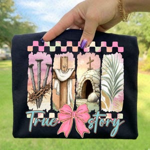 May include: Black t-shirt featuring a watercolor design with religious imagery. The design includes nails, a crown of thorns, a cross, a tomb, and palm leaves. A pink bow and the words "True Story" are also included in the design.