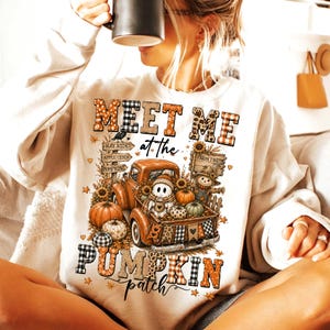 May include: A cream-colored crewneck sweatshirt featuring a graphic print with the text 'MEET ME at the PUMPKIN patch'. The design includes an orange truck carrying pumpkins, a ghost, and a scarecrow, surrounded by autumn-themed patterns.