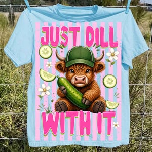 May include: Light blue t-shirt with a graphic of a Highland cow wearing a green cap and holding a cucumber. The text "JUST DILL WITH IT" is in pink. Cucumber slices and white flowers are also featured.