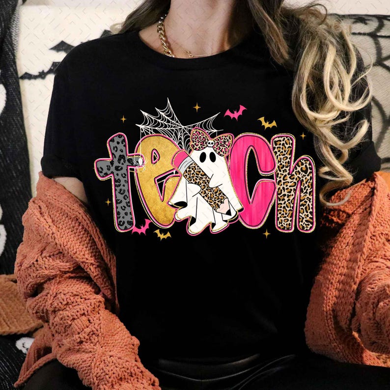 Peut inclure: Un t-shirt noir avec le mot TEACH &eacute;crit en grandes lettres color&eacute;es. La lettre A est remplac&eacute;e par un fant&ocirc;me blanc portant un n&oelig;ud &agrave; motif l&eacute;opard et tenant un crayon. Le motif inclut des toiles d'araign&eacute;e, des chauves-souris et des taches.
