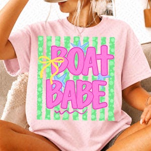 May include: A light pink t-shirt featuring a graphic design with the text BOAT BABE in bold pink letters. The background of the graphic consists of vertical green stripes with polka dots, accented by a yellow bow and light blue star shapes.