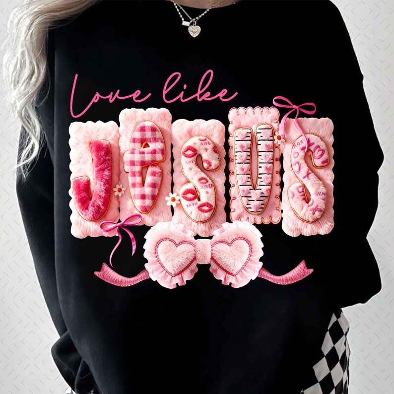 Op de afbeelding: Zwart sweatshirt met de tekst "Love like Jesus" in roze script. Het woord "Jesus" is weergegeven in roze, getextureerde letters met decoratieve elementen, waaronder lippen en harten. Een roze strik en een hartvormig ontwerp staan onderaan.