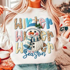 May include: White sweatshirt with a colorful embroidered design featuring a snowman and the words "Winter Winter Season". The snowman has a blue hat and scarf, an orange carrot nose, and black button eyes. The text is in various colors and fonts.