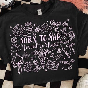 May include: Black t-shirt with a pink graphic design featuring medical symbols, flowers, bows, and the text "BORN TO YAP forced to chart." The design includes a stethoscope, bandages, and a nurse's shoe.