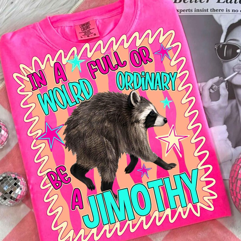 May include: A bright pink t-shirt featuring a central graphic of a raccoon. The design includes a wavy border, colorful stars, and the text: IN A WOLRD FULL OR ORDINARY BE A JIMOTHY. The shirt has a crew neckline and short sleeves with a visible brand label.