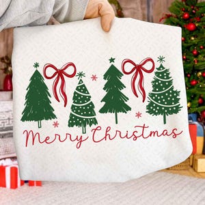 Puede incluir: Tela blanca con diseño navideño. Presenta árboles de Navidad verdes, lazos rojos y copos de nieve. Las palabras "Merry Christmas" están escritas en rojo en la parte inferior.