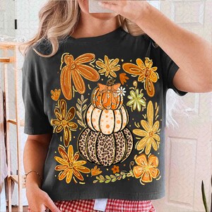 May include: Dark gray t-shirt featuring a stacked pumpkin design with a leopard print base, orange polka dots, and a bow. The shirt is adorned with orange and brown floral accents, perfect for autumn or fall.