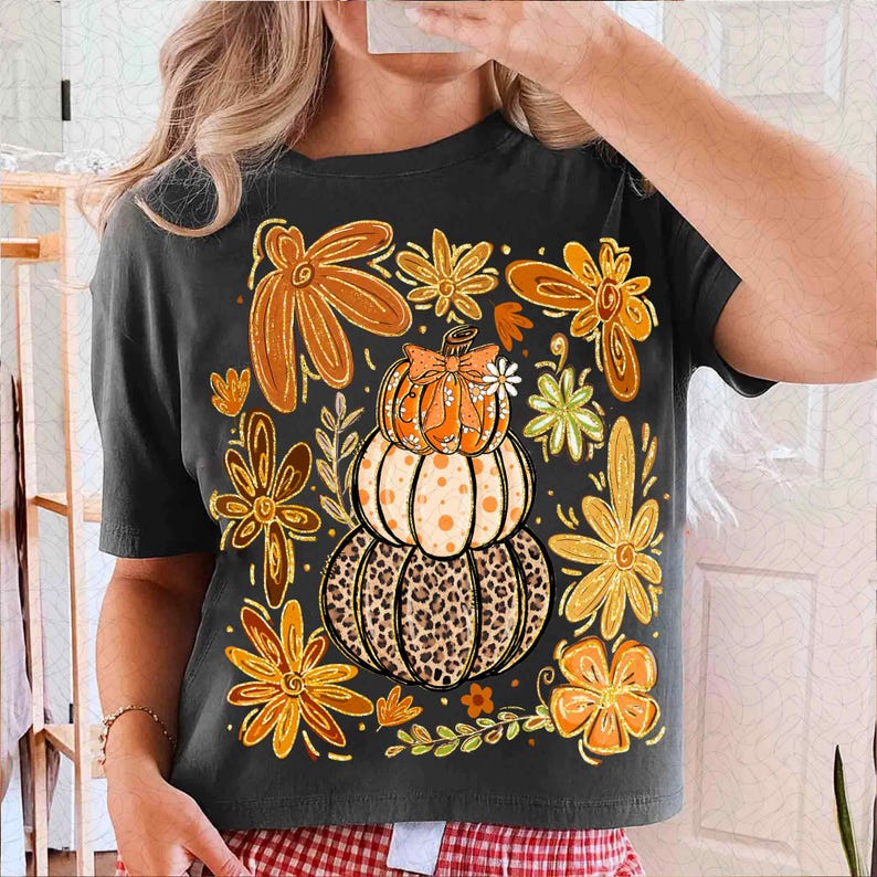 May include: Dark gray t-shirt featuring a stacked pumpkin design with a leopard print base, orange polka dots, and a bow. The shirt is adorned with orange and brown floral accents, perfect for autumn or fall.