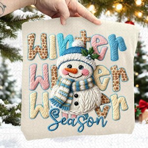 May include: Embroidered pillow with a snowman design. The pillow features the words "Winter Winter Season" in various colors and patterns. The snowman has a blue hat and scarf, an orange carrot nose, and black button eyes. The pillow is cream-colored.