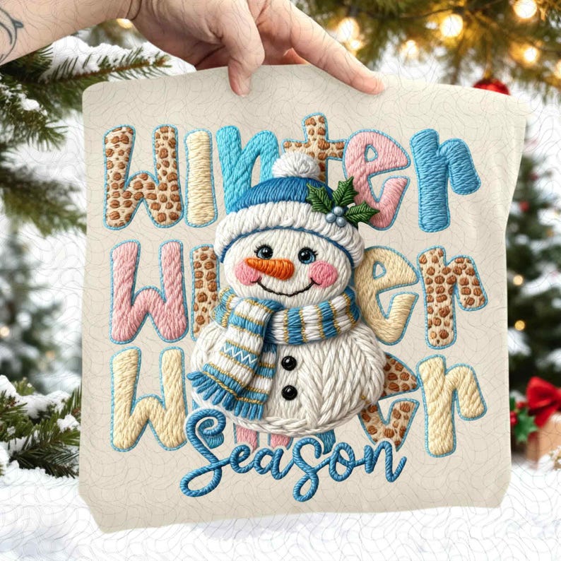 May include: Embroidered pillow with a snowman design. The pillow features the words "Winter Winter Season" in various colors and patterns. The snowman has a blue hat and scarf, an orange carrot nose, and black button eyes. The pillow is cream-colored.
