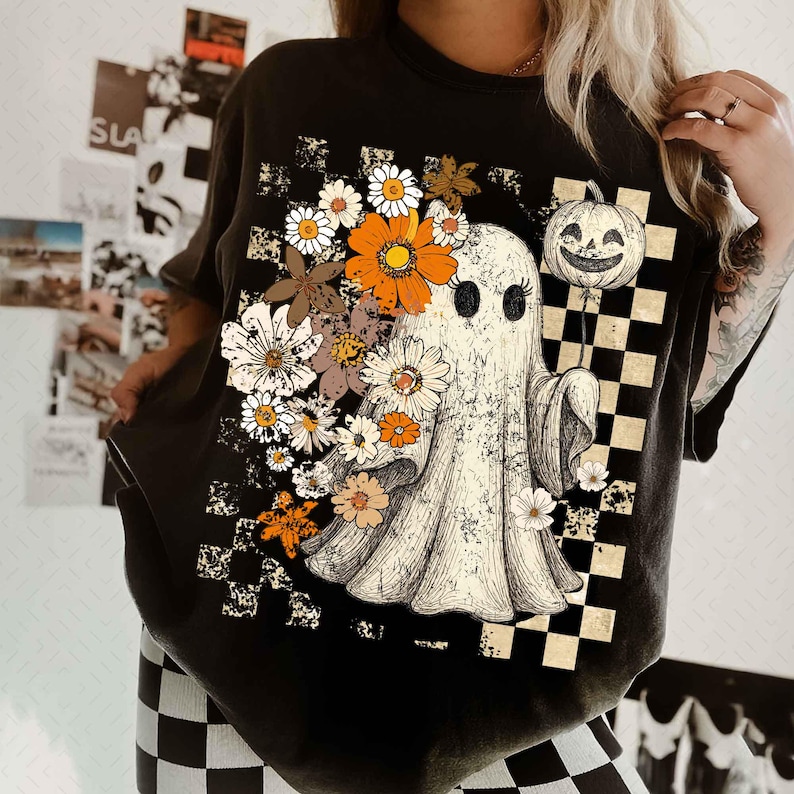May include: Black t-shirt featuring a vintage-style graphic of a ghost holding a pumpkin balloon, surrounded by flowers. The design includes a checkered pattern and a distressed look, perfect for Halloween or fall.