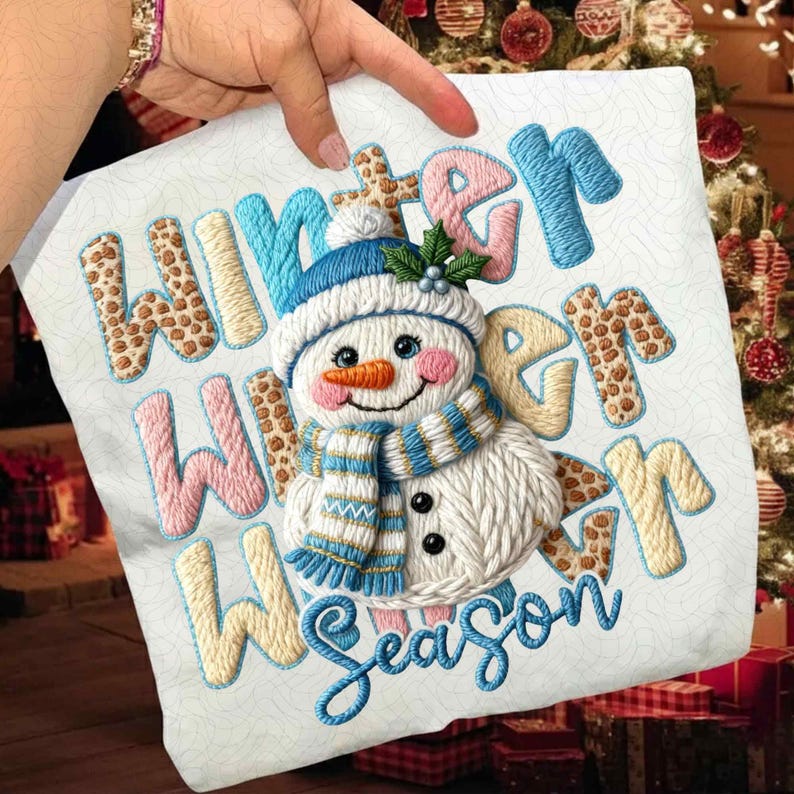 May include: White fabric with embroidered text and a snowman design. The text reads "Winter Season" in blue, pink, and tan colors. The snowman has a blue hat, scarf, and an orange carrot nose.