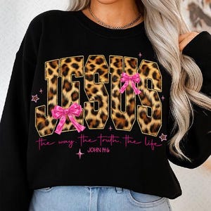May include: Black sweatshirt featuring the word "JESUS" in a leopard print design, adorned with pink bows and star shapes. The script text below reads "the way. the truth. the life" and "JOHN 14:6" in pink.
