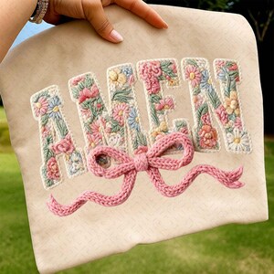May include: A beige fabric item with the word "AMEN" embroidered in a floral design. The letters are filled with pink, blue, and yellow flowers and green leaves. A pink knitted bow is attached below the letters.