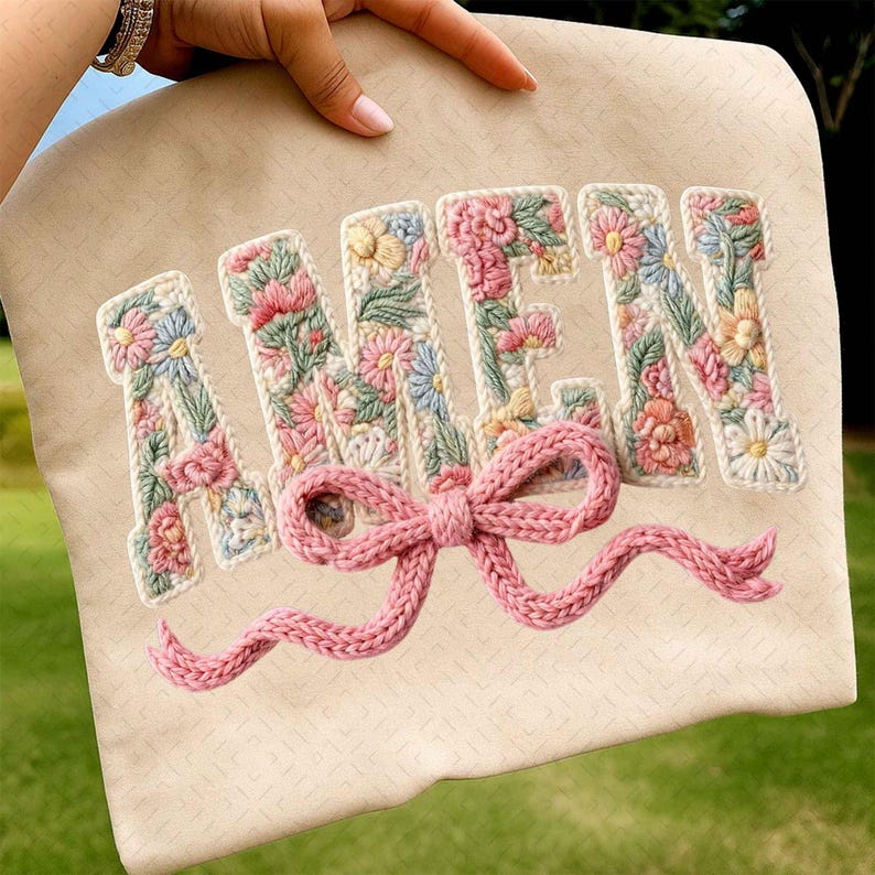May include: A beige fabric item with the word "AMEN" embroidered in a floral design. The letters are filled with pink, blue, and yellow flowers and green leaves. A pink knitted bow is attached below the letters.