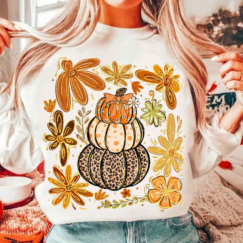 May include: White sweatshirt featuring a stacked pumpkin design with a leopard print base, orange polka dots, and a bow. Surrounding the pumpkin are various orange and gold floral elements, creating a fall-themed aesthetic.