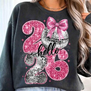 May include: Dark gray sweatshirt featuring a sparkly pink "26" design with a disco ball and pink bow. The word "hello" is written in cursive. Pink star accents complete the design.