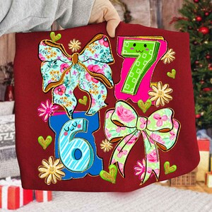 May include: A red fabric item featuring the numbers 6 and 7, along with two floral bows and decorative hearts and flowers. The bows have floral patterns in pink, blue, and green. The numbers are outlined in blue and pink.