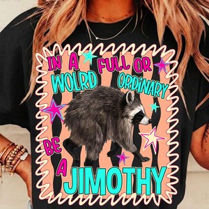 May include: A black t-shirt featuring a graphic print of a raccoon centered on a peach-colored background with a wavy border. The text reads IN A FULL OR WOLRD ORDINARY BE A JIMOTHY in bright blue letters, accented by pink and blue stars.