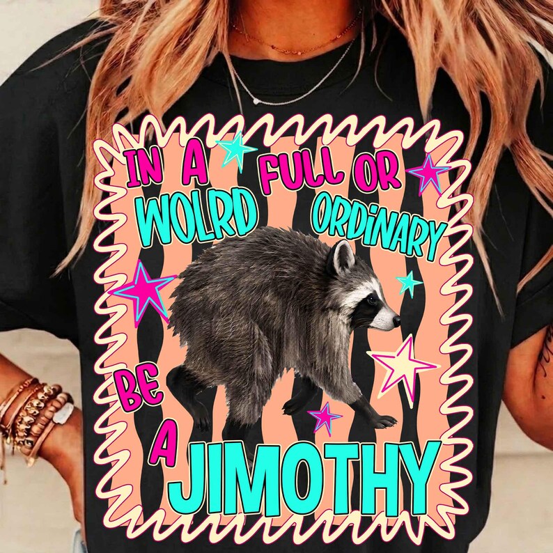 May include: A black t-shirt featuring a graphic print of a raccoon centered on a peach-colored background with a wavy border. The text reads IN A FULL OR WOLRD ORDINARY BE A JIMOTHY in bright blue letters, accented by pink and blue stars.