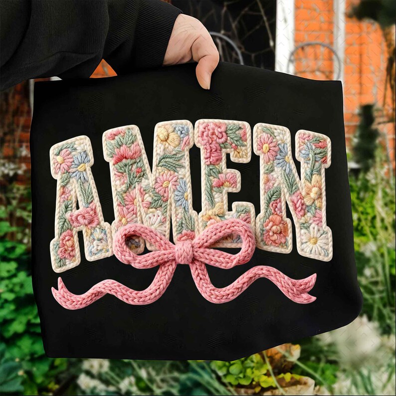May include: Black tote bag with the word "AMEN" embroidered in floral letters. A pink knitted bow is centered below the letters. The bag is held by a person.
