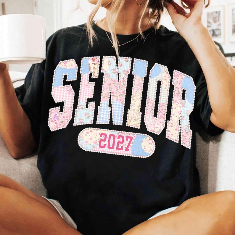 Peut inclure: T-shirt noir avec le mot "SENIOR" dans un motif patchwork de motifs floraux et vichy. L'ann&eacute;e "2027" est affich&eacute;e sur un patch ovale. Le t-shirt est de style d&eacute;contract&eacute;.