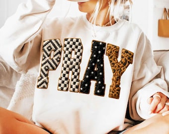 May include: A white sweatshirt with the word "PRAY" in a patchwork design. The letter "P" is black and white, the "R" is black with white stars, the "A" is black and white, and the "Y" is brown.