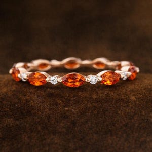 Könnte beinhalten: Ein roségoldener Ring mit abwechselnd orangefarbenen, marquiseförmigen Edelsteinen und kleinen weißen, runden Edelsteinen. Der Ring ist vor einem dunkelbraunen, strukturierten Hintergrund platziert.