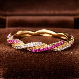 May include: A gold-toned ring with a twisted design, featuring alternating rows of small, sparkling white and pink gemstones. The ring is presented in a brown velvet jewelry box, showcasing its elegant design.