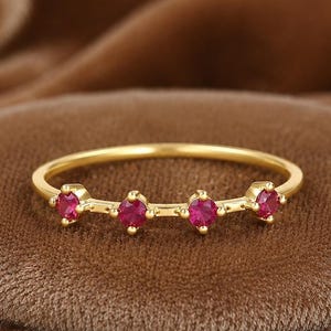 Op de afbeelding: Een delicate gouden ring met vier kleine, levendige rode edelstenen. De ring heeft een dunne band en de edelstenen zijn gelijkmatig verdeeld. Gepresenteerd op een bruin, getextureerd oppervlak, wat het elegante ontwerp benadrukt.