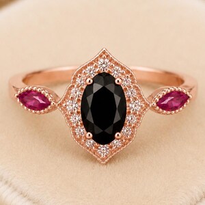 May include: A rose gold ring featuring a central oval-cut black stone surrounded by a halo of small white stones. The setting has a pointed, scalloped edge and includes a marquise-cut red stone on each side of the band, creating a detailed design.