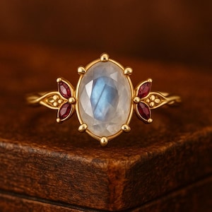 May include: A gold ring featuring an oval, iridescent moonstone center stone. Two clusters of three small, red gemstones flank the center stone. The ring is set on a wooden surface.