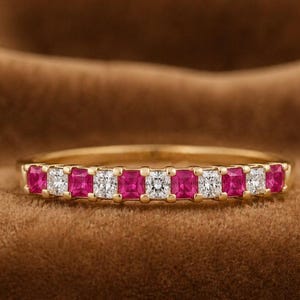 Op de afbeelding: Een gouden ring met een band bezet met afwisselend vierkant geslepen roze edelstenen en heldere diamanten. De ring is geplaatst tegen een bruine achtergrond, die de levendige kleuren van de edelstenen benadrukt. De ring is een sieraad.